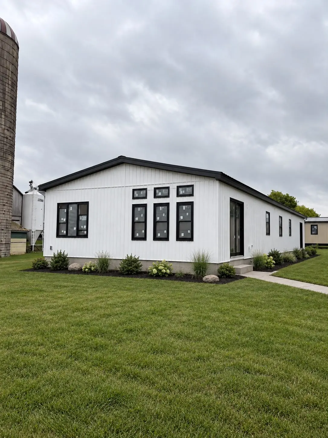 A newly finished home with white board and batten siding and black window frames, standing on a lawn beside a farm silo with the window labels still in place.