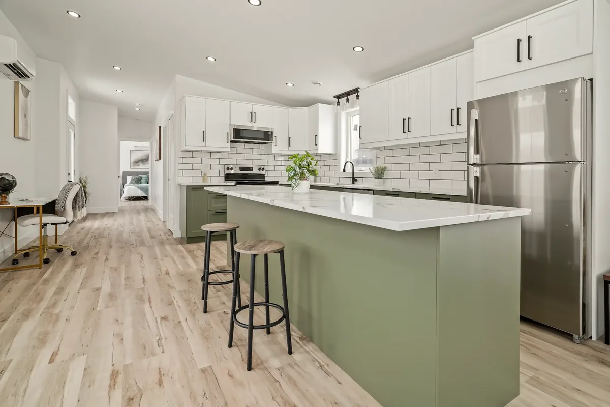 The kitchen of a finished REDI-HOME, looking down the length of the house: a quartz-topped island with stools, sage green lower cabinets under white uppers, a white subway tile backsplash, and vinyl plank flooring running through to the bedroom door.